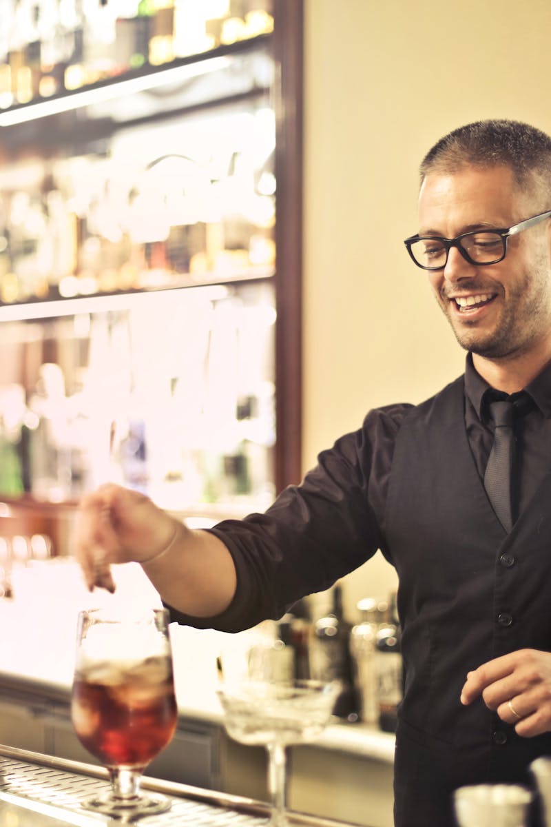 Home Happy young male barkeeper standing at counter and preparing alcohol cocktail for order while working in modern pub