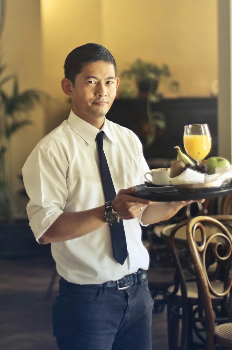 Home Confident young ethnic waiter in elegant clothes holding tray with food and drinks and looking at camera while serving tables in stylish restaurant
